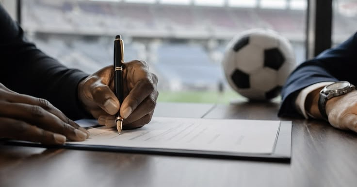 Manos firmando un contrato junto a una pelota de fútbol, con un estadio de fondo